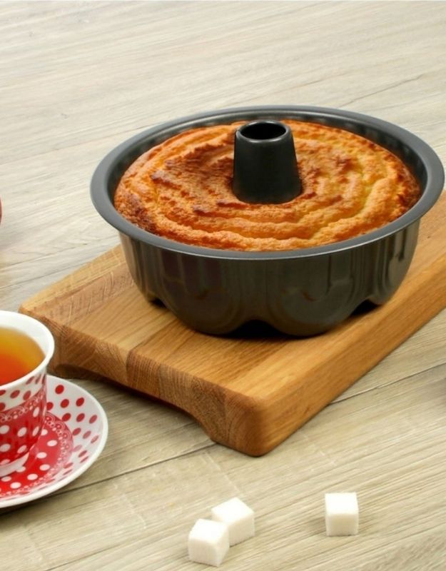 Bundt cake in a pan on a wooden board with a cup of tea and sugar cubes on a light wooden surface. Freshly baked cake in black nonstick bundt cake pan on wooden board.