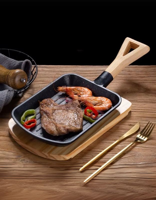 A black carbon steel cast iron skillet with a wooden handle and tray, containing cooked meat and vegetables, served on a wooden table with gold-colored cutlery beside it.