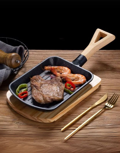 A black carbon steel cast iron skillet with a wooden handle and tray, containing cooked meat and vegetables, served on a wooden table with gold-colored cutlery beside it.