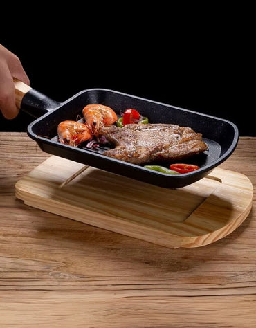 Small black frying pan with cooked food on a wooden stand. Nonstick grill pan with wooden handle lifted from serving tray, showing steak and shrimp inside.