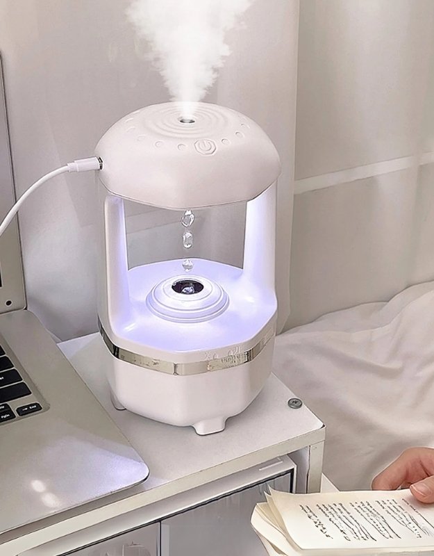 A white USB aroma diffuser with a visible water tank, emitting mist, placed on a table next to a person reading a book.