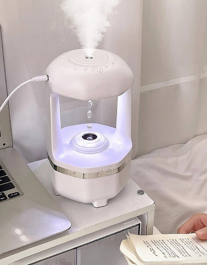 A white USB aroma diffuser with a visible water tank, emitting mist, placed on a table next to a person reading a book.