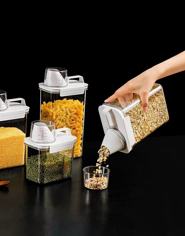 Container with a pouring spout being used to pour pasta into a glass on a dark surface.
Hand pouring cereal from airtight food dispenser container into measuring cup.