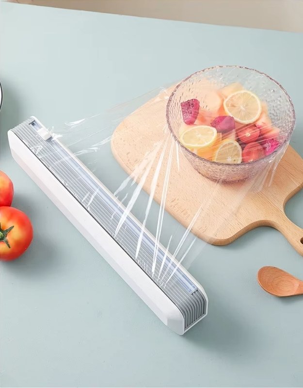 A white magnetic plastic cling wrap dispenser with a slide cutter, shown mounted on a wooden board in a kitchen setting with fruits and vegetables in the background.