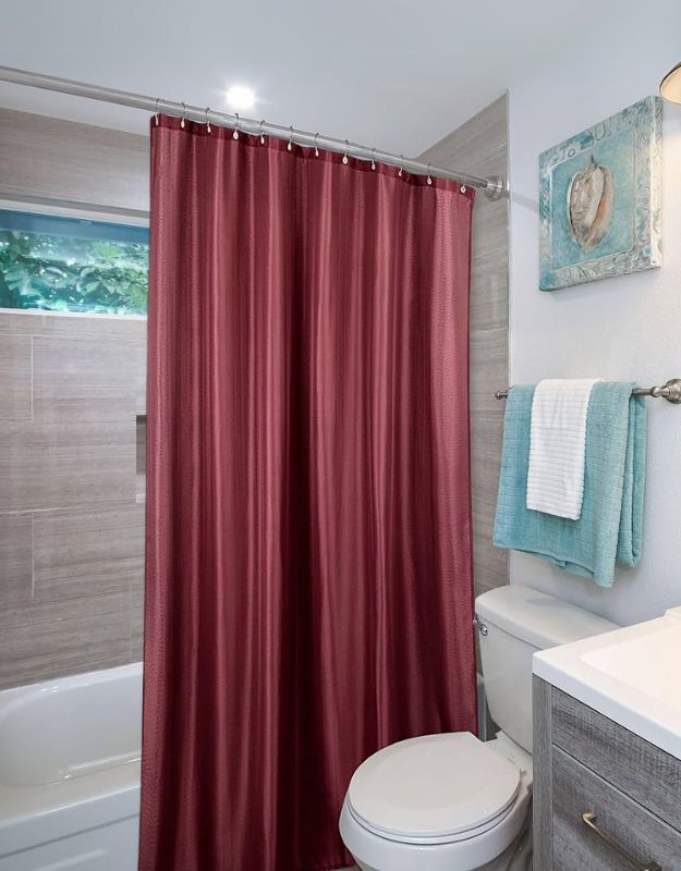 A red polyester bathroom curtain with embroidery, hanging in a bathroom with a light fixture above and towels on the right side.
