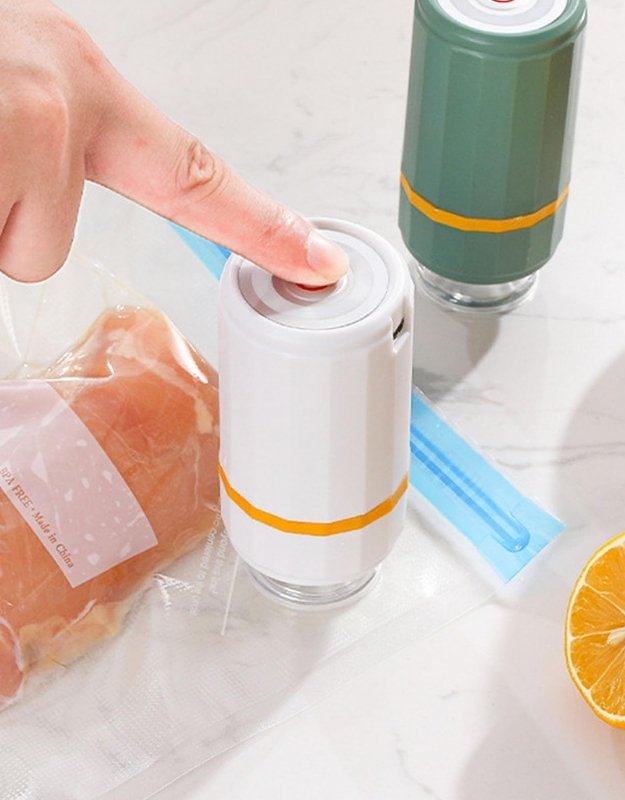 The image shows a Mini Vacuum Sealer in white and green colors, with a hand pressing a button on the top. In the background, there are vacuum-sealed bags with food items visible and a lemon.
