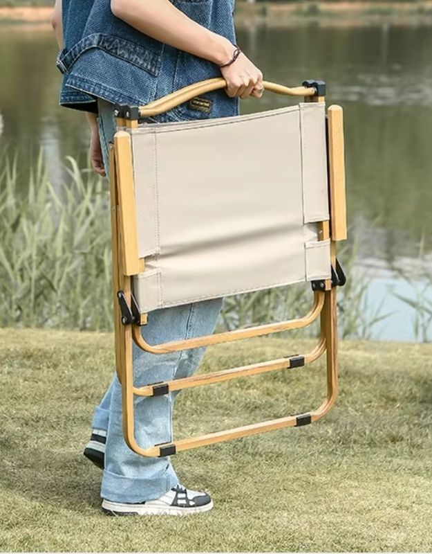 A person carrying a foldable camping chair. The chair has a lightweight wooden or bamboo frame and beige fabric seating, designed for portability and outdoor use such as picnics, camping, or fishing. The person is holding it by a built-in handle, and the background features grass and a lake, indicating a natural outdoor setting.