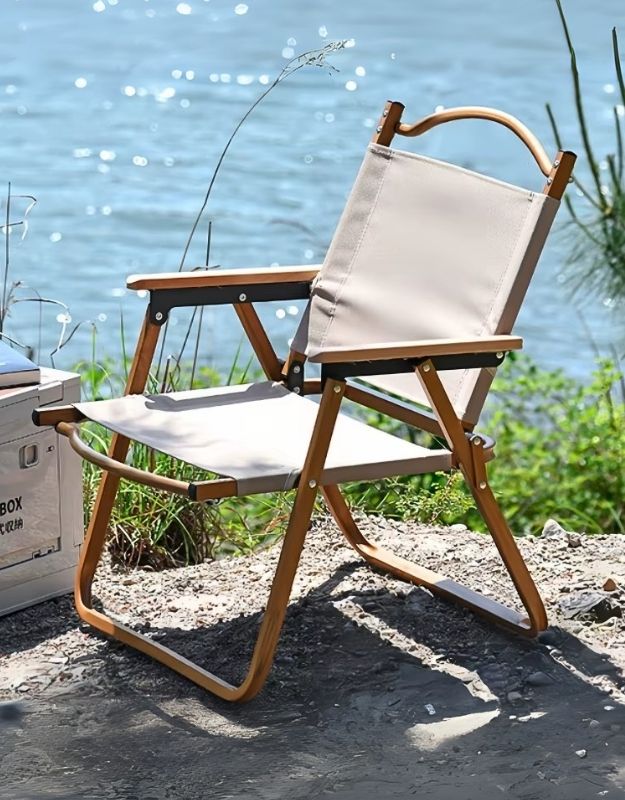 A portable folding chair with a beige fabric seat and wooden frame, set outdoors with a body of water in the background.