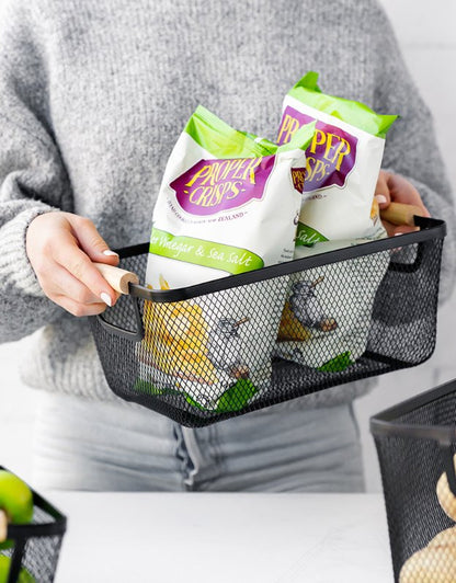 Rectangular black mesh basket with wooden handles storing packets of crisps.