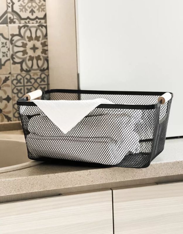 Rectangular black mesh basket with wooden handles used for storing rolled towels in kitchen or bathroom.