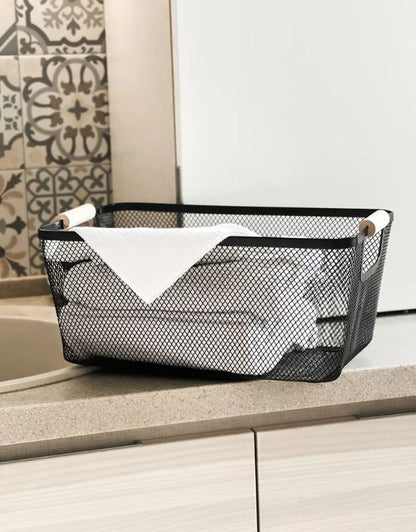 Rectangular black mesh basket with wooden handles used for storing rolled towels in kitchen or bathroom.
