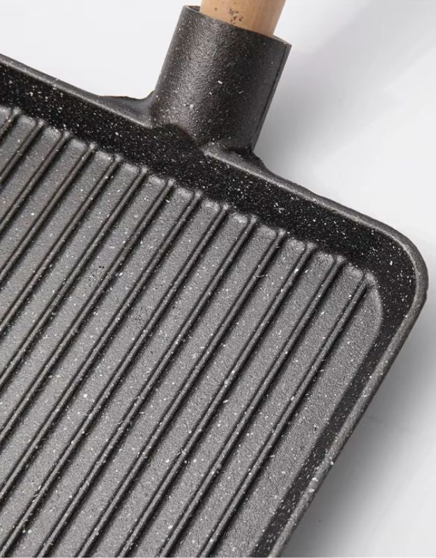 Close-up of a black grill pan with a wooden handle on a white background. Close-up of rectangular nonstick grill pan with ribbed surface for grilling.
