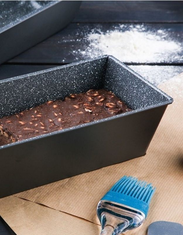 Baked brownie in a loaf pan with flour on a wooden surface and a blue brush. Rectangular non-stick loaf pan with chocolate nut cake batter inside.