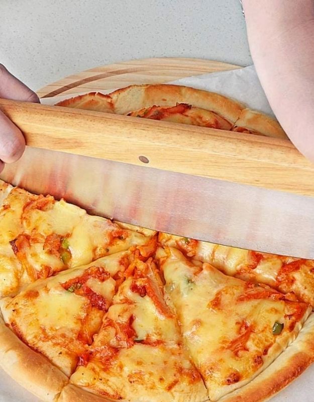 Person using a wooden pizza cutter to slice a pizza on a white surface. Close-up of rocker pizza cutter slicing a cheese pizza with vegetables.