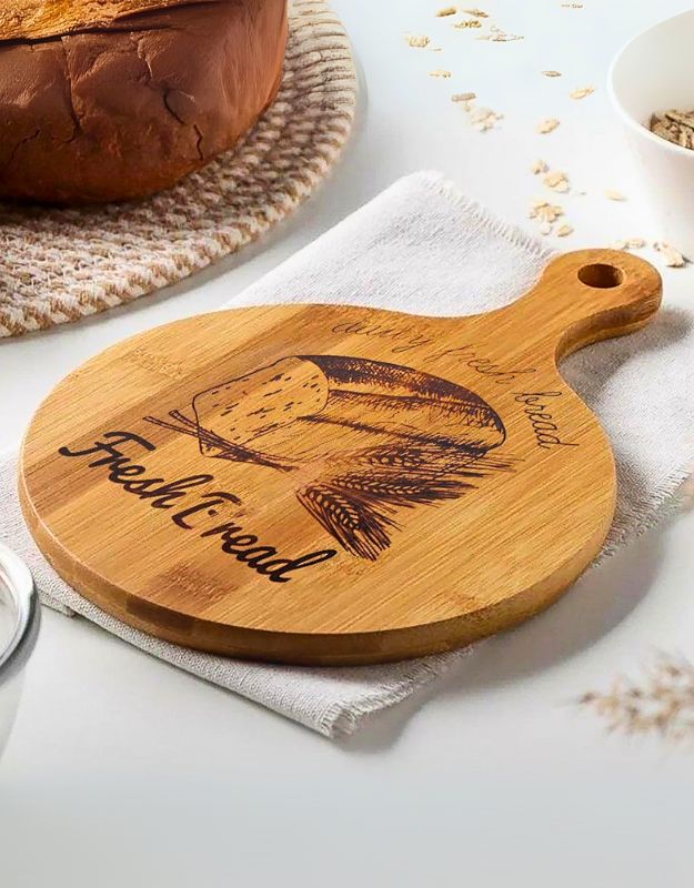 A round bamboo wood cutting board with a handle, featuring the text 'Fresh Bread' and a bread loaf image engraved on it, resting on a white surface with a loaf of bread in the background.