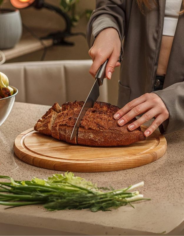 Person slicing a loaf of bread on a wooden cutting board in a kitchen 
setting. Cutting fresh bread on round wooden chopping board in kitchen.