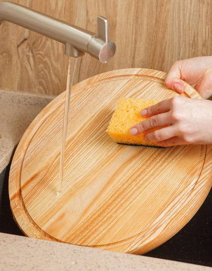 Person cleaning a wooden cutting board with a sponge under running 
water. Washing round wooden chopping board under running water