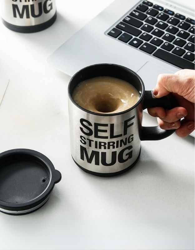 A stainless-steel silver self-stirring mug with black and blue accents on a desk next to a computer keyboard, with a person's hand holding the mug.