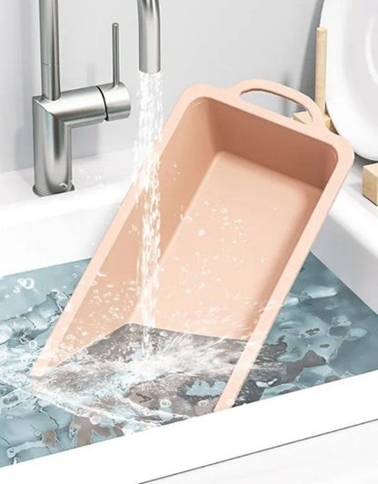 Pink cleaning brush being rinsed under running water in a sink. Nonstick silicone loaf pan being washed under running water.