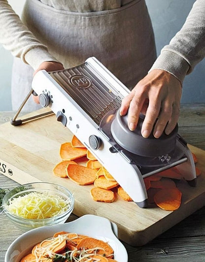 Person using a mandoline slicer to slice sweet potatoes on a wooden cutting board. Adjustable mandoline slicer slicing sweet potatoes with hand guard.