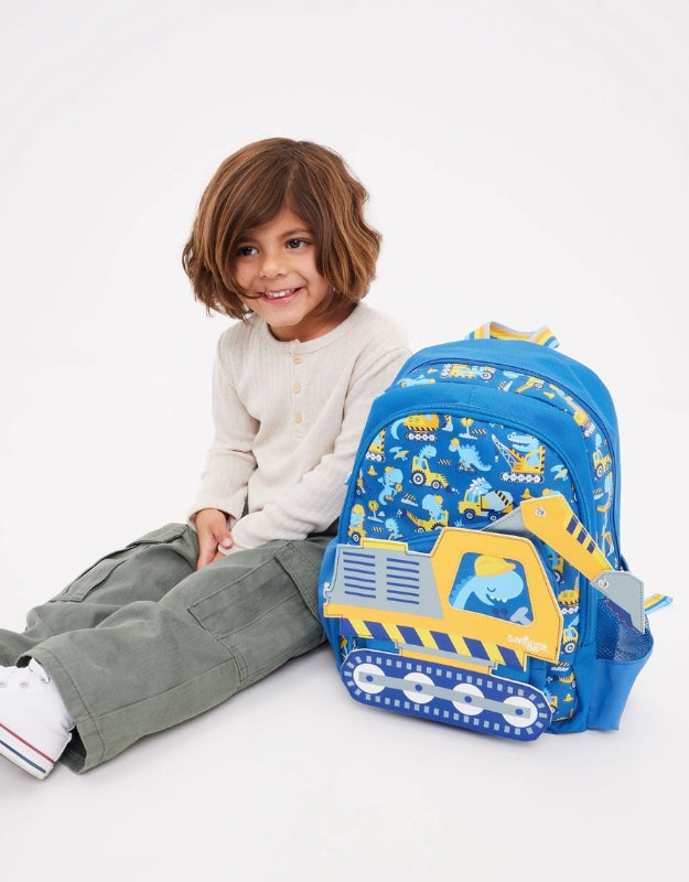 Smiling child sitting beside the Smiggle Big Dreams Junior Character Backpack with blue and yellow construction theme.