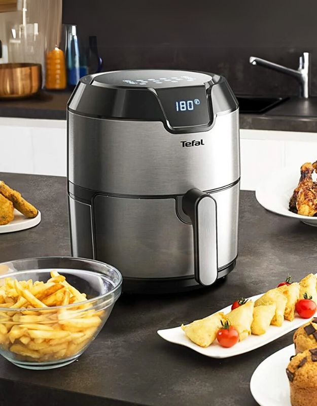 A Tefal Easy Fry Oil Less 4.2L Air Fryer with digital interface displayed on a kitchen counter surrounded by plates of food including fries and chocolate muffins.