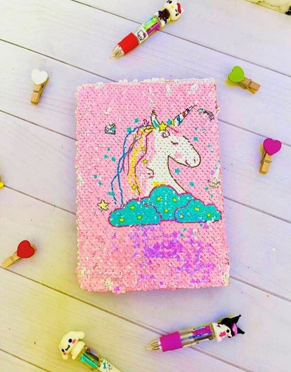 Pink notebook with unicorn design on a wooden surface with stationery items.
