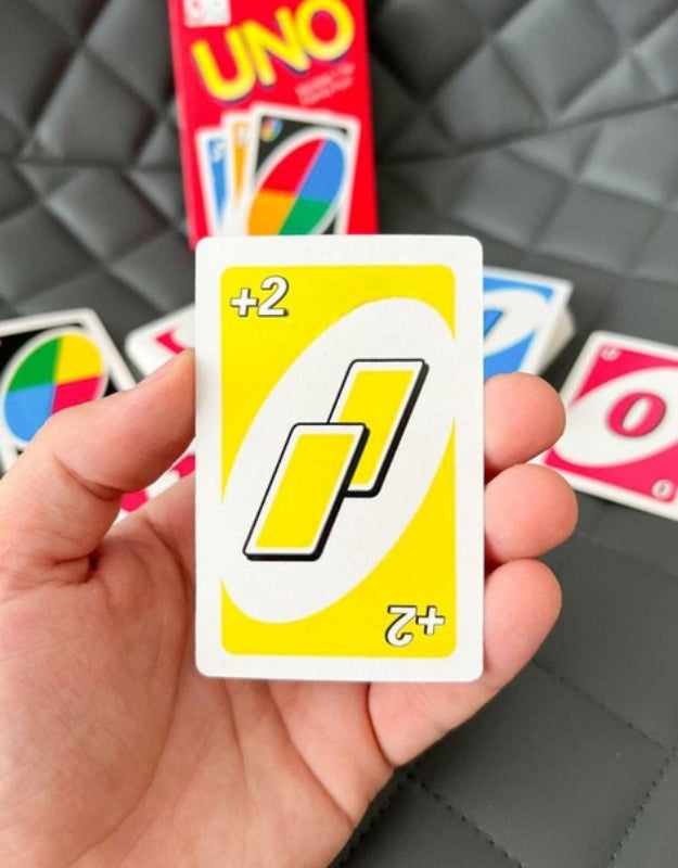 A close-up shot of a hand holding a single yellow +2 UNO card, with the game box blurred in the background
