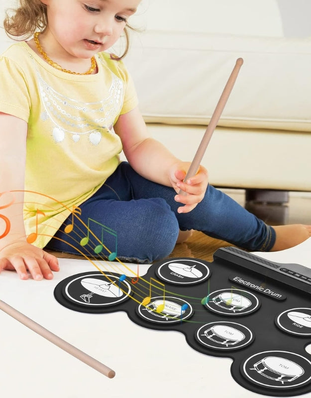 An angled view of the portable electronic drum kit showing the drum pad and its connected foot pedals and a pair of wooden drumsticks