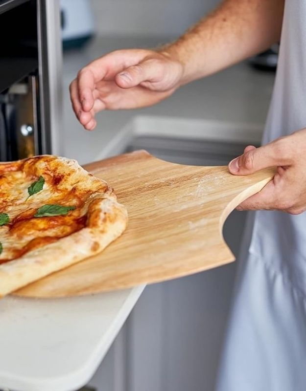 Person holding a wooden pizza peel with a pizza on it, likely in a kitchen setting. Person using a wooden pizza peel to slide a pizza into the oven.